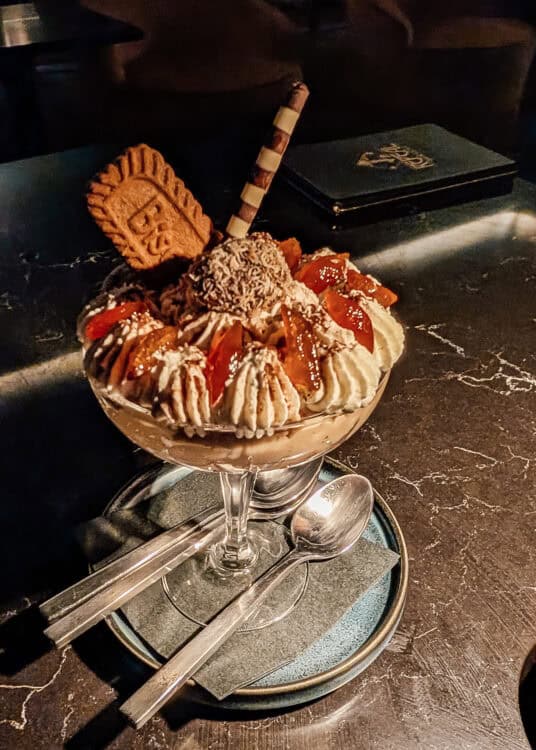 A decadent chocolate dessert is served in a glass sundae dish topped with piped whipped cream, chocolate truffles, caramelized fruit, a striped wafer stick, and a Biscoff cookie. The dessert sits on a dark marble table with two spoons, creating an elegant presentation in a dimly lit restaurant.