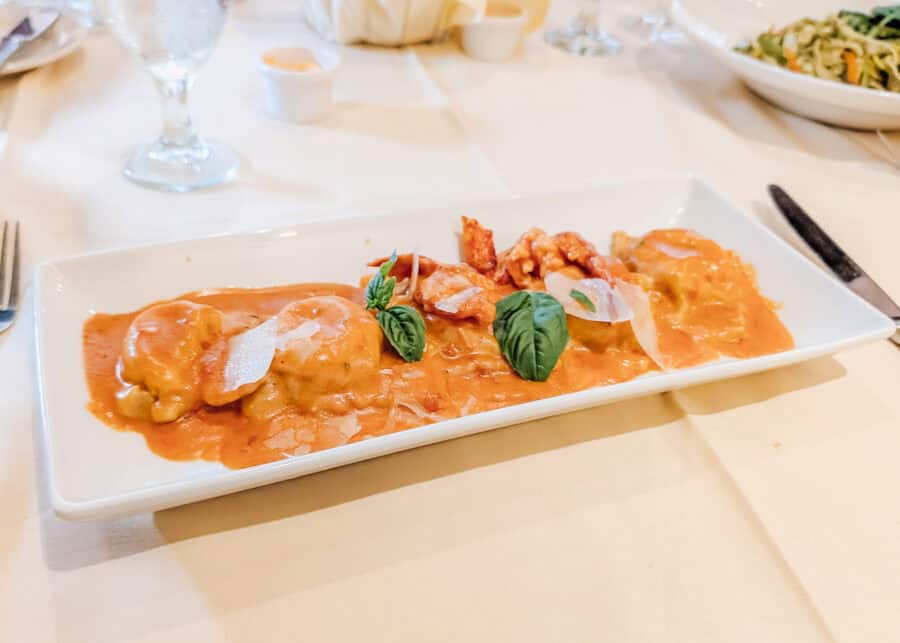 A rectangular white plate holds lobster ravioli covered in a creamy tomato sauce and topped with fresh basil leaves, shaved Parmesan, and chunks of lobster. The pasta is served on a white tablecloth with water glasses and a side salad in the background, creating an elegant restaurant dining scene.