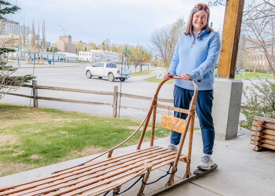 A smiling woman stands on the runners of a wooden dog sled and holds the handle in downtown Anchorage. The traditional sled offers a close up look at Alaska dog mushing culture in the city.