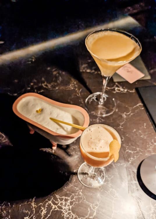 Three craft cocktails are arranged on a dark marble table in a dimly lit bar, including a creamy martini, a foamy cocktail garnished with an orange peel, and a drink served in a miniature bathtub with a small gold spoon. The creative presentation and moody lighting create an upscale cocktail lounge atmosphere.