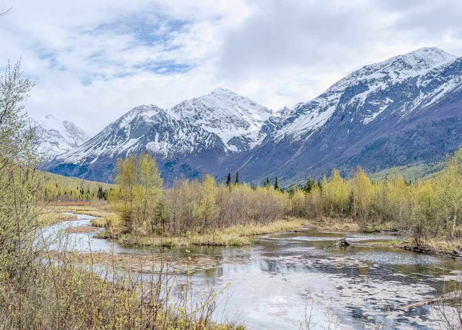 A winding river flows through a forest of fresh spring greenery beneath towering snow covered mountains in Alaska. The peaceful water and rugged peaks showcase the dramatic natural scenery surrounding Anchorage.