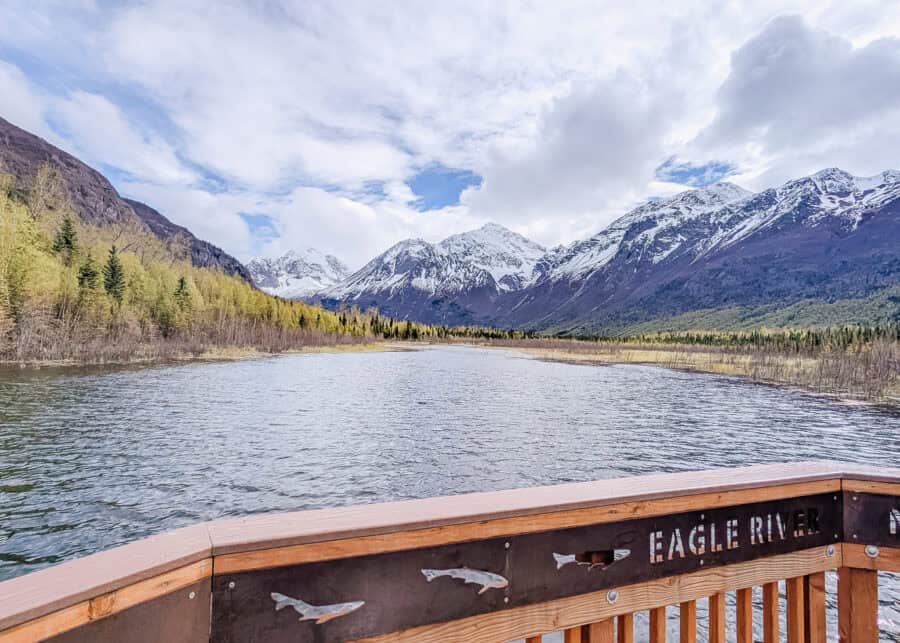 Eagle River flows through a broad valley surrounded by forest and dramatic snowcapped mountains in Alaska. A wooden viewing deck in the foreground is marked "EAGLE RIVER" and decorated with metal fish silhouettes.