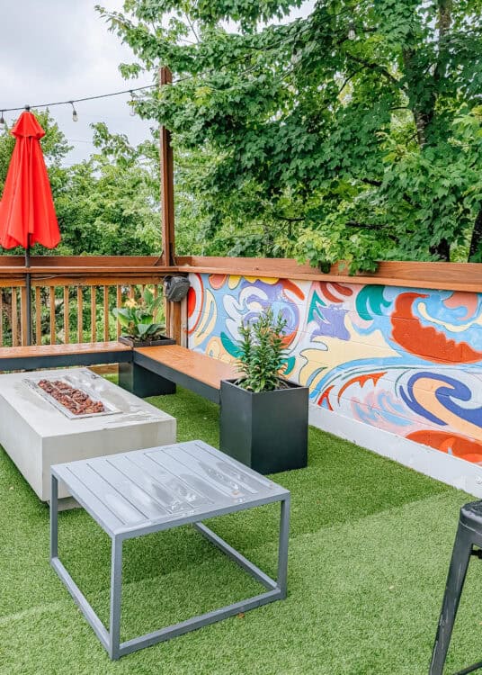 An outdoor patio features a modern fire table, built in wooden bench seating, potted plants, and a colorful abstract mural along one wall. Artificial turf, string lights, and a bright red patio umbrella create a relaxed gathering space surrounded by leafy trees.