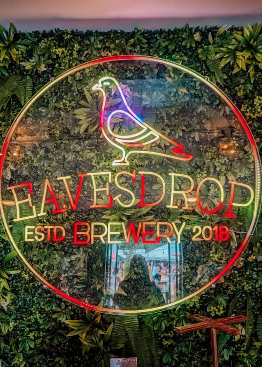 A large circular neon sign with a pigeon logo glows against a lush green plant wall inside Eavesdrop Brewery. The illuminated sign reads "EAVESDROP ESTD BREWERY 2018," creating a colorful focal point and popular photo backdrop inside the brewery.