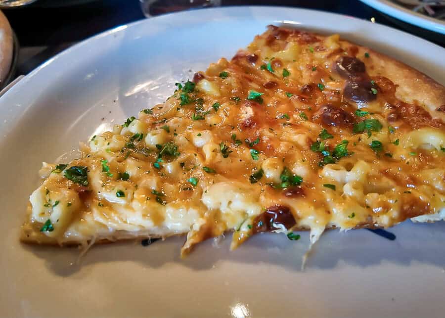 A slice of pizza topped with creamy mac and cheese melted cheese and fresh parsley on a white plate. The close view highlights the rich cheesy macaroni topping and golden crust.