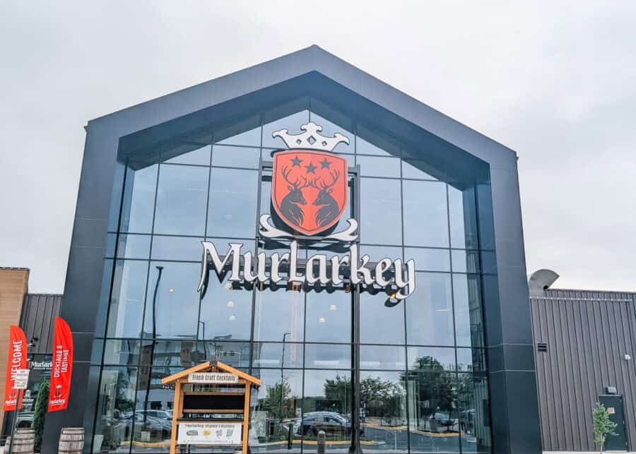 The modern glass entrance to Murlarkey Distilled Spirits features the distillery's large logo with two deer beneath a crown displayed prominently on the front of the building. Red welcome flags and a wooden stand labeled "Fresh Craft Cocktails" greet visitors outside the contemporary distillery under an overcast sky.