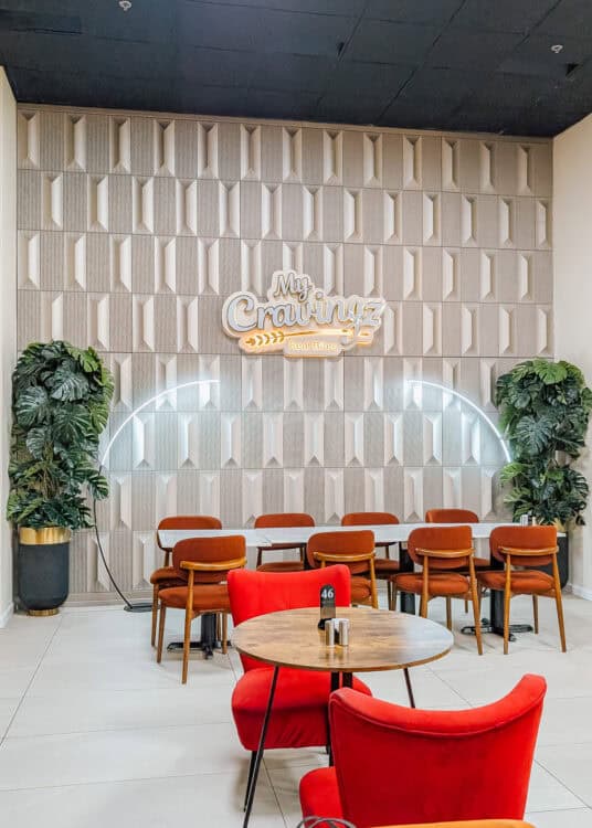 A modern restaurant dining area features red velvet chairs, wooden tables, large potted plants, and a textured accent wall with a glowing sign that reads "My Cravingz Real Bites." A curved LED light frames the seating area, creating a stylish and inviting space for guests.