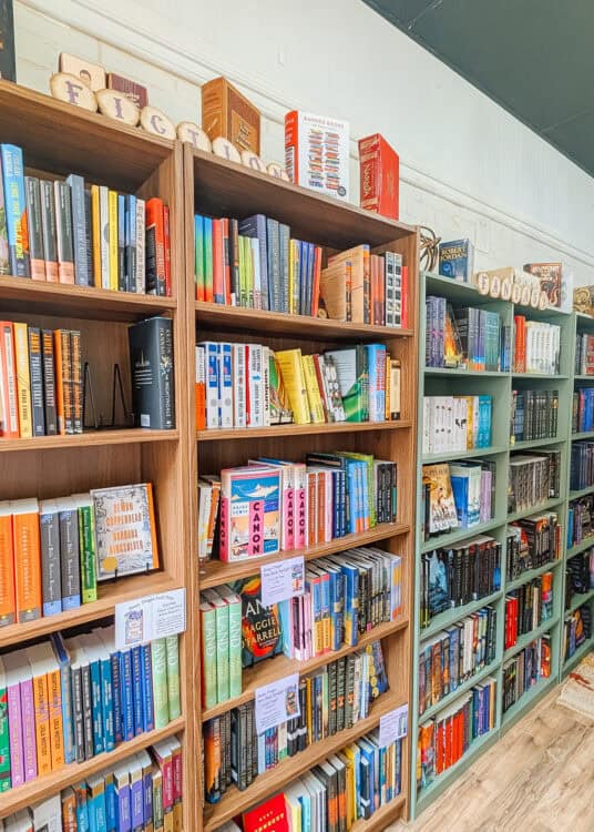 Tall wooden bookshelves filled with colorful novels line the walls of an independent bookstore, with sections labeled "Fiction" and "Fantasy" displayed across the top. Featured book displays and neatly organized shelves create a cozy browsing space with white brick walls and wood flooring.