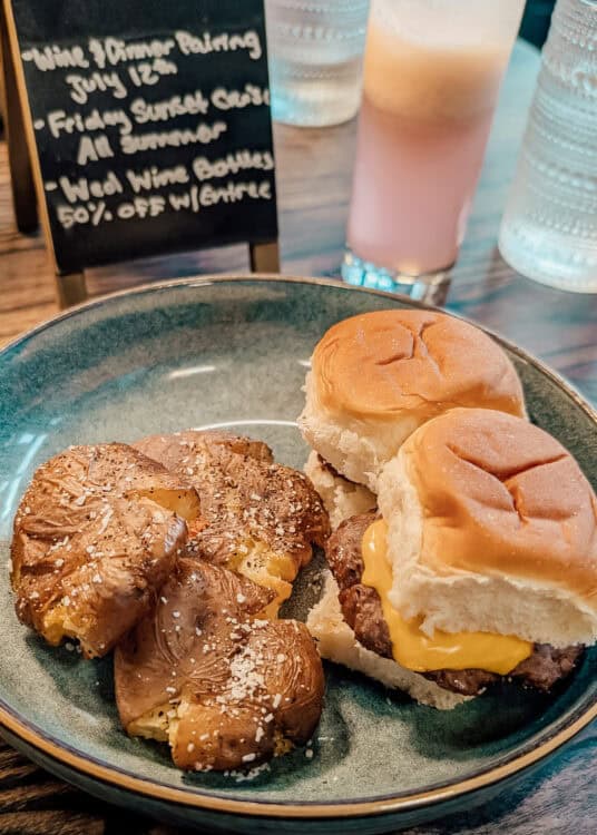 Two cheeseburger sliders on soft buns are served with roasted baby potatoes sprinkled with herbs and coarse salt on a blue plate. A pink layered cocktail and a chalkboard in the background advertise specials including "Wine & Dinner Pairing July 12," "Friday Sunset Cocktails All Summer," and "Wed Wine Bottles 50% Off w/ Entree," creating a casual restaurant dining scene.
