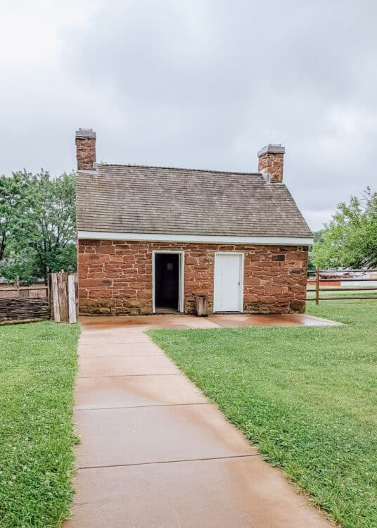 A small stone building that served as enslaved quarters stands at the end of a wet concrete walkway surrounded by a grassy lawn beneath an overcast sky. The structure features thick stone walls, two chimneys, a steep shingled roof, and simple doorways, illustrating the basic living conditions provided for enslaved people at the historic site.