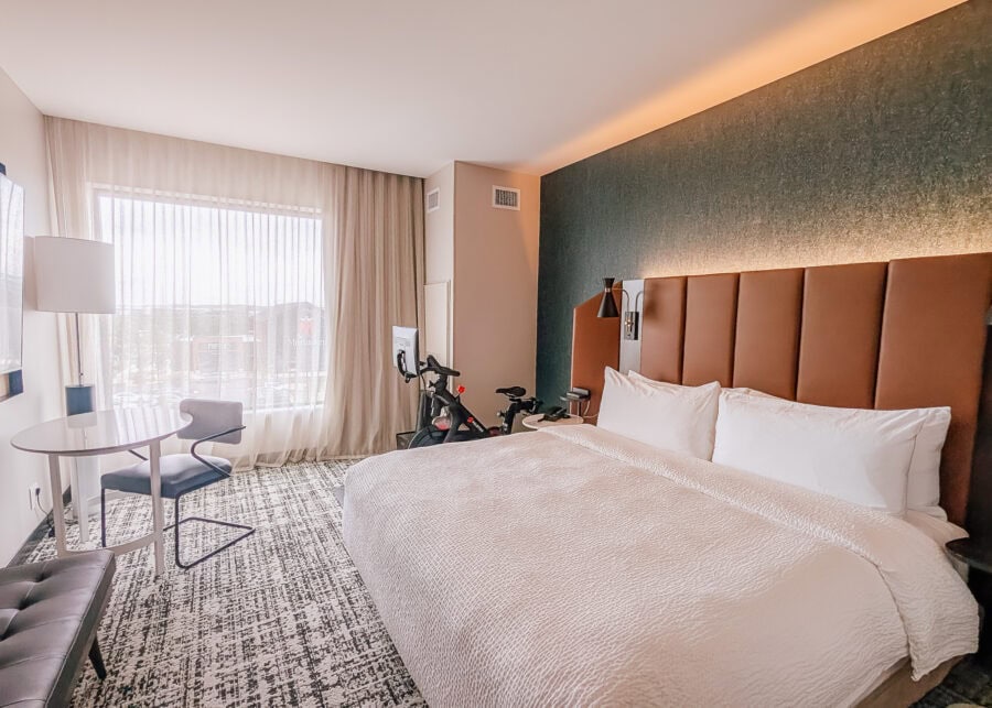 A modern hotel room features a king bed with a tall upholstered headboard, a small table and chair by a large window, and a stationary exercise bike positioned beside the bed. Neutral decor, floor to ceiling curtains, and natural light create a bright, comfortable space designed for both relaxation and in room workouts.