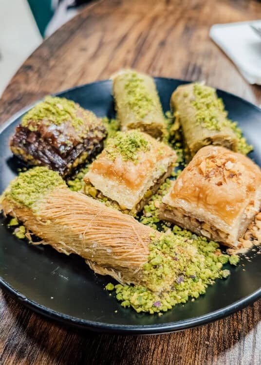A black dessert plate is filled with assorted Middle Eastern pastries, including baklava, pistachio rolls, chocolate baklava, and shredded phyllo pastry, all generously topped with crushed pistachios. The colorful assortment of flaky, syrupy sweets is arranged on a wooden table, showcasing a variety of traditional desserts in one serving.