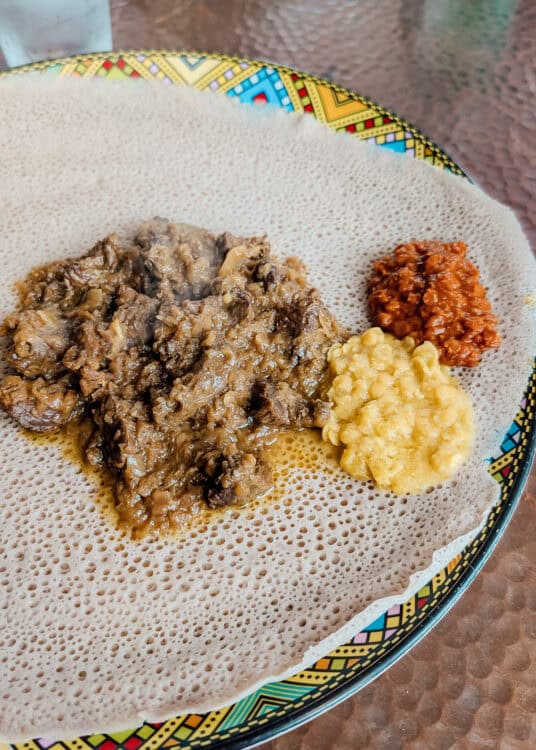 An Ethiopian meal is served on soft injera bread with a generous portion of tender beef stew alongside creamy yellow lentils and a scoop of bright red chili sauce. The colorful patterned plate sits on a restaurant table, highlighting the traditional presentation and contrasting textures of the meal.