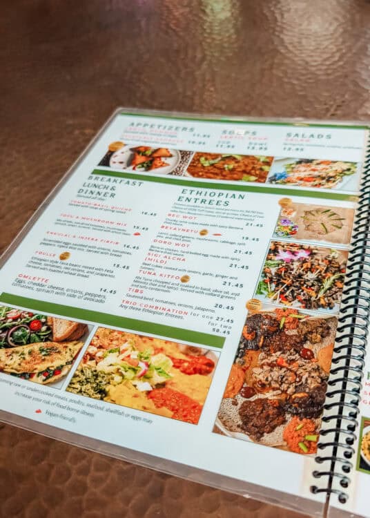 An open spiral bound restaurant menu rests on a textured tabletop, showing colorful photos of Ethiopian dishes alongside breakfast, lunch, dinner, appetizer, soup, and salad options. Visible menu text includes the headings "APPETIZERS," "SOUPS," "SALADS," "BREAKFAST LUNCH & DINNER," and "ETHIOPIAN ENTREES," with featured dishes such as "Tomato Basil Quiche," "Tofu & Mushroom Mix," "Enkulal & Injera Firfir," "Foulle," "Omlette," "Beyaynetu," "Doro Wot," "Mild Alicha," "Tuna Kitfo," "Tibs," and "Trio Combination," along with their listed prices.