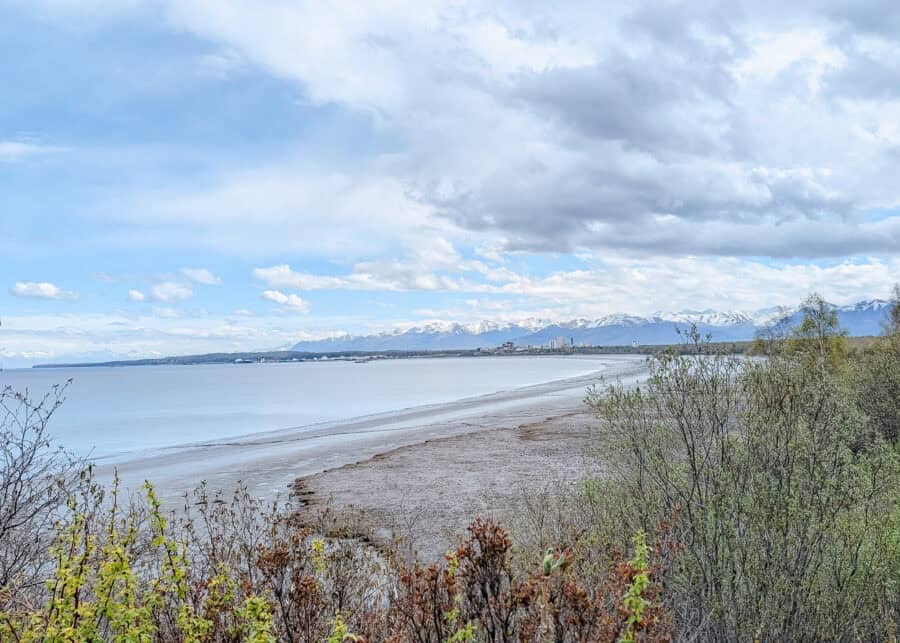 View across the calm waters and tidal flats of Cook Inlet toward downtown Anchorage. Snowcapped Chugach Mountains stretch behind the city beneath dramatic clouds and patches of blue sky.