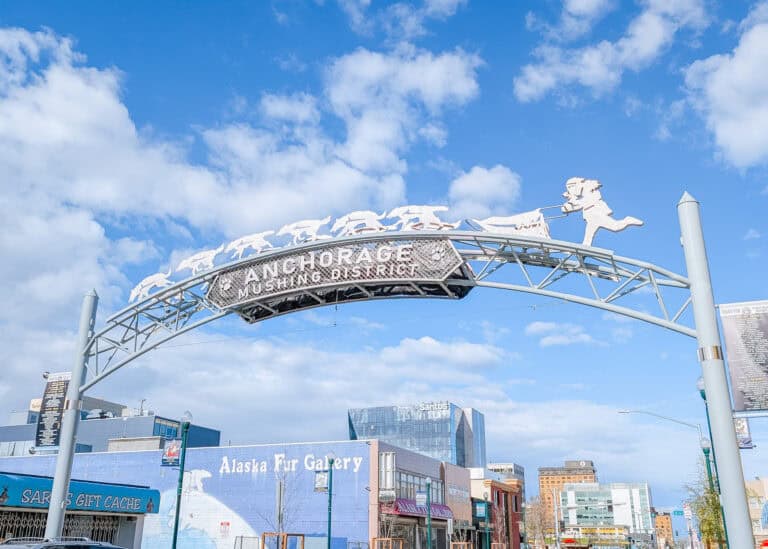 The "ANCHORAGE MUSHING DISTRICT" arch spans a downtown Anchorage street with metal silhouettes of a musher and sled dogs racing across the top. The Alaska Fur Gallery and other storefronts sit below under a bright blue sky with scattered clouds.
