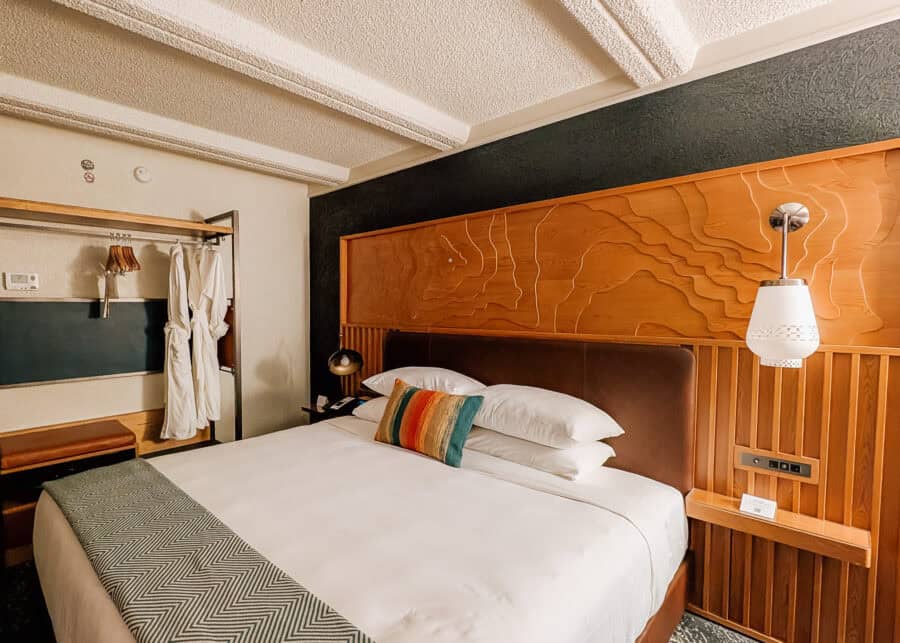 A cozy hotel room with a king bed framed by a warm wood headboard carved with topographic lines. White robes hang on an open wardrobe beside the bed with a colorful striped pillow and modern pendant light adding color and warmth.