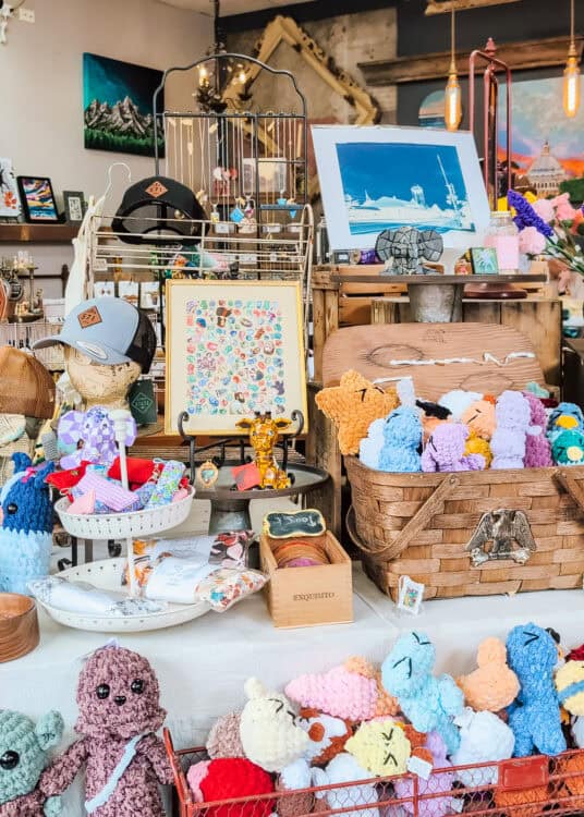 A colorful gift shop display is filled with handmade crocheted plush animals, artisan jewelry, locally designed hats, framed artwork, and small handcrafted gifts arranged on wooden crates and baskets. The eclectic boutique features rustic decor, hanging pendant lights, and shelves showcasing work from local artists and makers.