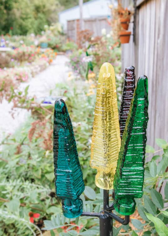 our colorful textured glass garden sculptures in teal, yellow, dark purple, and green stand among leafy plants. A flower filled garden and wooden fence create a softly blurred background behind the decorative glass pieces.