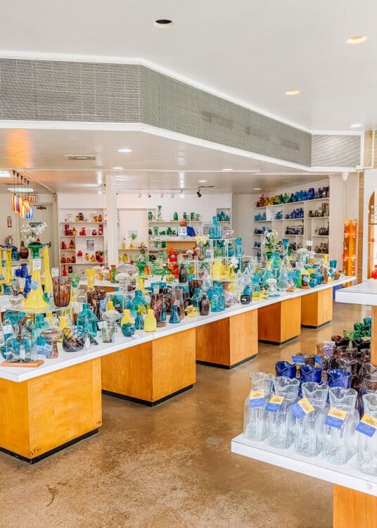 A bright glass shop is filled with colorful hand blown glass vases, pitchers, bottles, and decorative pieces displayed across wooden tables and wall shelves. The glassware is arranged in vivid shades of blue, green, yellow, red, purple, and clear glass throughout the shop.