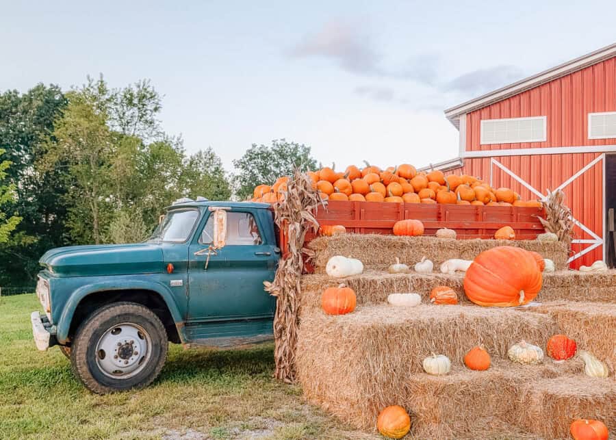 A vintage teal farm truck is parked beside stacked hay bales covered with orange and white pumpkins and gourds. The truck bed is overflowing with pumpkins in front of a large red barn.