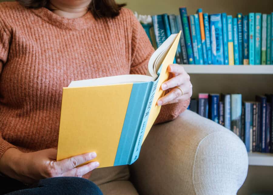 A woman in a rust colored sweater sits in an armchair reading a yellow hardcover book. Shelves filled with books arranged in shades of blue create a colorful backdrop for the cozy reading scene.
