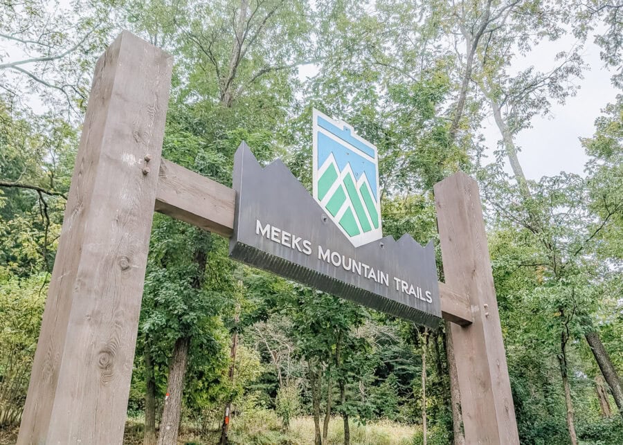 A wooden entrance sign reading "Meeks Mountain Trails" stands beneath a canopy of leafy green trees. A blue and green mountain logo tops the sign at the entrance to the wooded trail system.