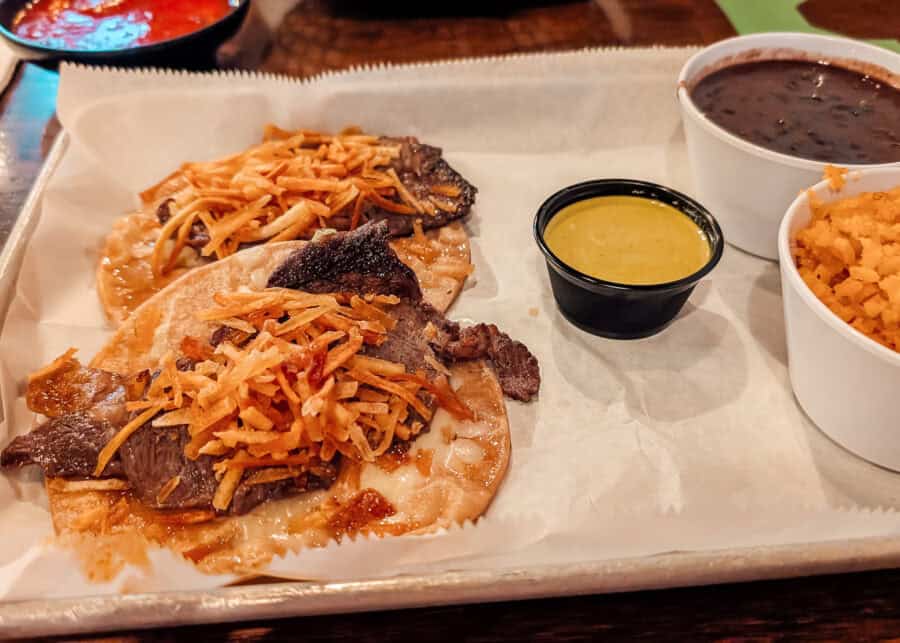 Two steak tacos topped with crispy potatoes on a paper lined tray. Black beans. Mexican rice. And a cup of green sauce are served alongside the tacos.