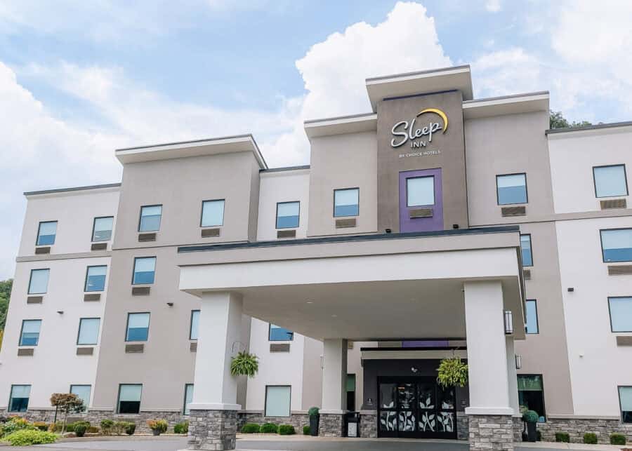 The exterior of a Sleep Inn by Choice Hotels features a modern light gray facade with purple accents and a covered entrance. The four story hotel sits beneath a bright blue sky with large white clouds.