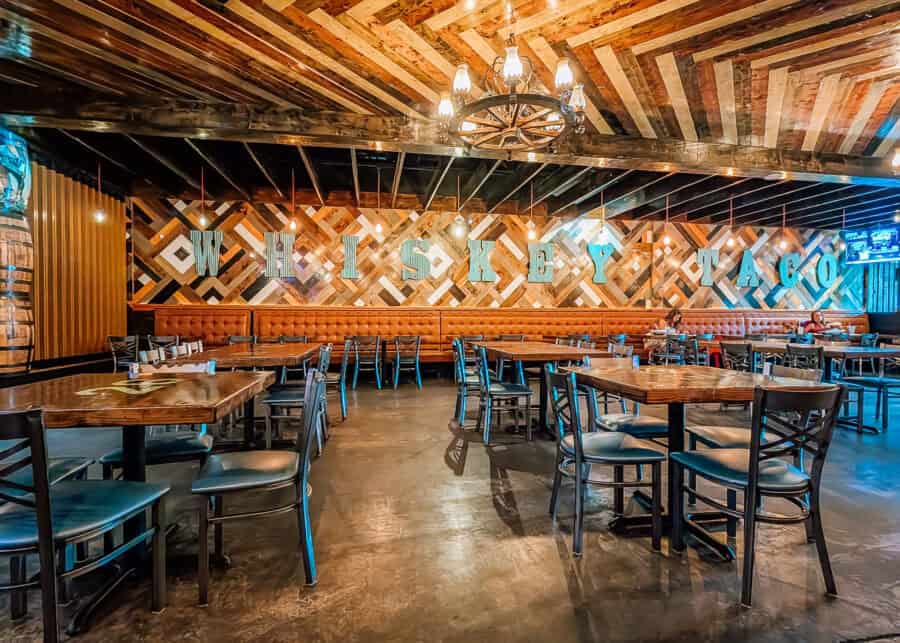 Rustic restaurant dining room with wooden tables and black chairs beneath warm pendant lights and a wagon wheel chandelier. Large turquoise letters spelling "WHISKEY TACO" stretch across a geometric wood accent wall above a long brown booth.