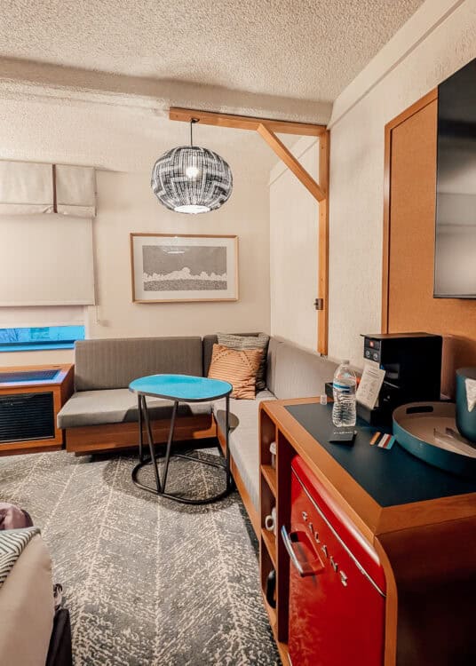 Hotel room sitting area with a gray corner sofa and small blue table beneath a woven pendant light. A coffee station and bright red mini fridge sit beside the seating area.