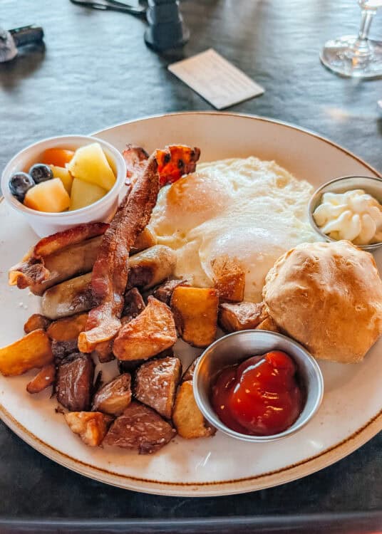 Breakfast plate with two fried eggs. Bacon. Sausage. Breakfast potatoes. A biscuit. Fresh melon and blueberries. Ketchup and a creamy sauce.