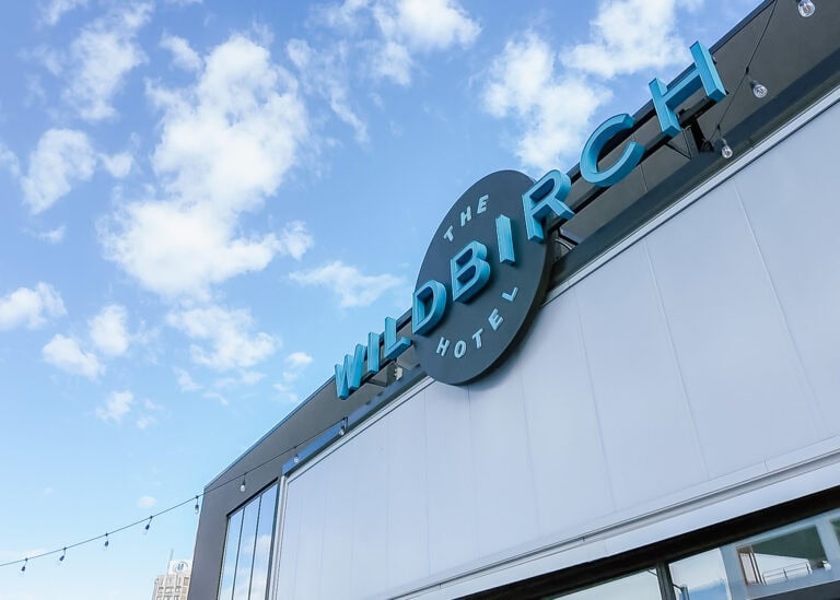 Exterior sign for "The Wildbirch Hotel" in large blue letters along the roofline. The modern hotel building is photographed from below against a bright blue sky with scattered white clouds.