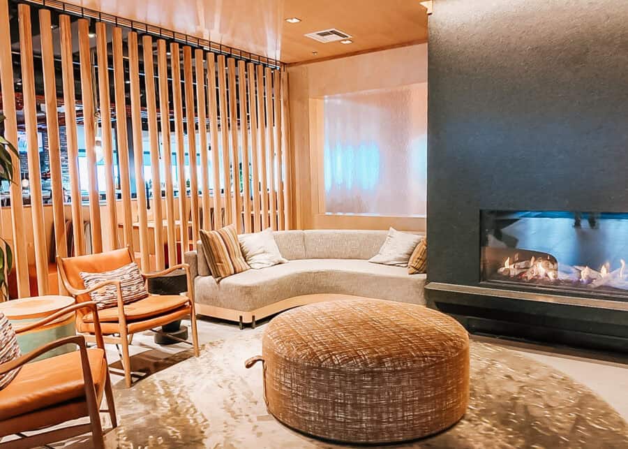 Cozy modern lounge with a curved cream sofa leather chairs and a large round ottoman arranged beside a glowing fireplace. Vertical wood slats and warm lighting create a relaxed contemporary seating area.