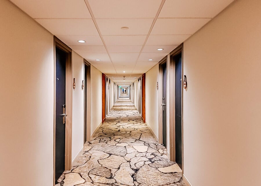 Long hotel hallway with dark guest room doors on both sides and patterned carpet resembling cracked stone. Recessed ceiling lights create a symmetrical view stretching toward the far end of the corridor.