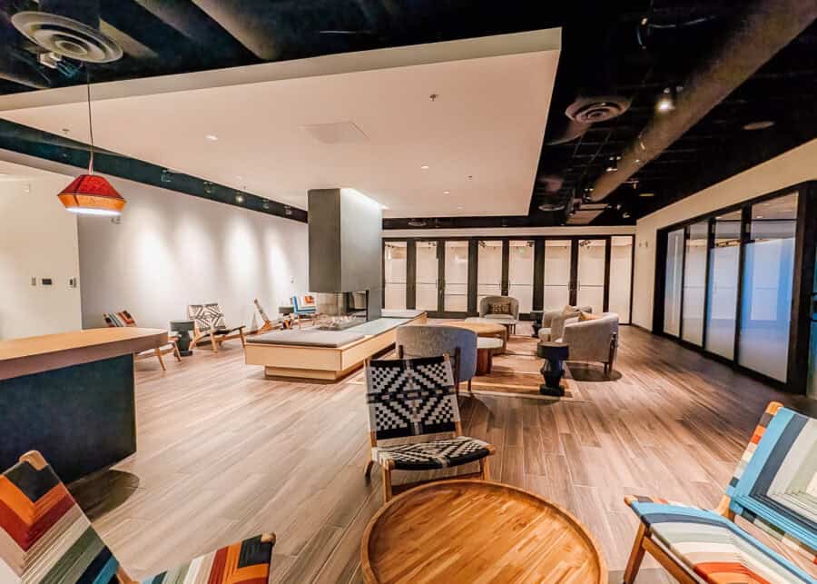 Spacious modern hotel lounge with colorful woven chairs arranged around wooden tables and sofas beside a central fireplace. Warm wood floors and geometric ceiling panels give the communal seating area a contemporary feel.