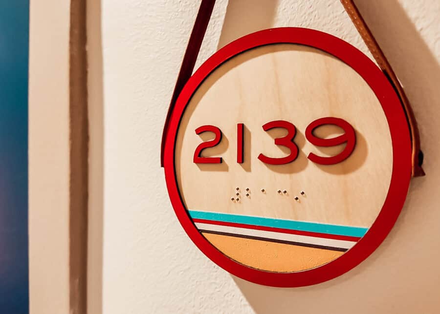 Round hotel room sign with the number 2139 in raised red lettering and Braille below. Teal. Red. White. And gold stripes decorate the bottom of the wood sign.
