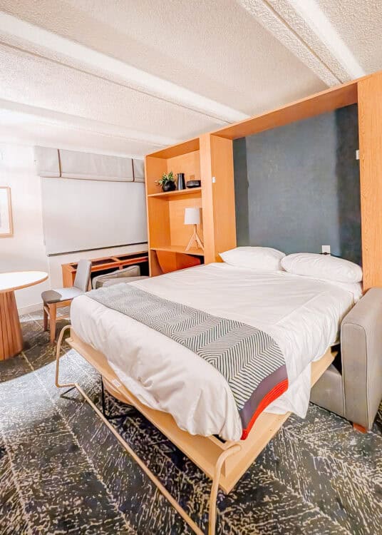 Hotel room with a queen size Murphy bed pulled down over the sofa and dressed in white bedding with a patterned runner. A desk and chair sit beside the bed with warm wood built ins framing the sleeping area.