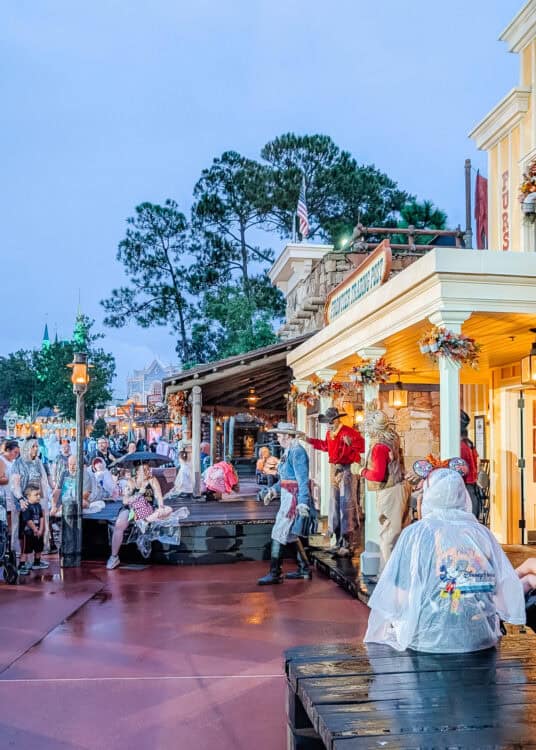 Performers dressed as frontier townspeople interact with guests outside the Country Bear Jamboree building in Frontierland at Magic Kingdom in Walt Disney World. Visitors wearing rain ponchos gather along the wet walkway at dusk while warm lights illuminate the rustic storefronts decorated with autumn garlands.