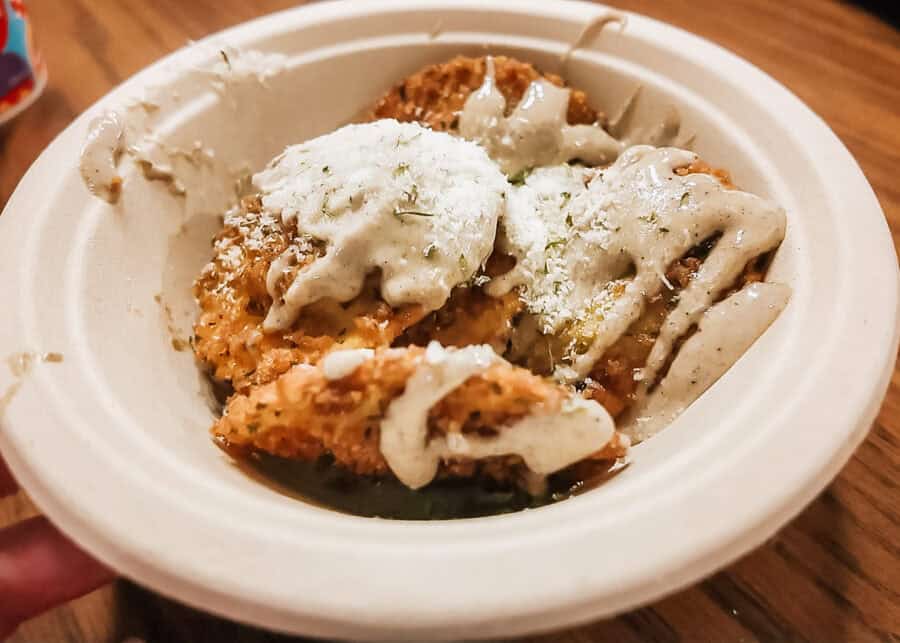 Several golden brown breaded and fried bites are served in a white bowl with a generous drizzle of creamy sauce. The crispy pieces are topped with grated cheese and a sprinkling of green herbs, creating a savory snack or appetizer.