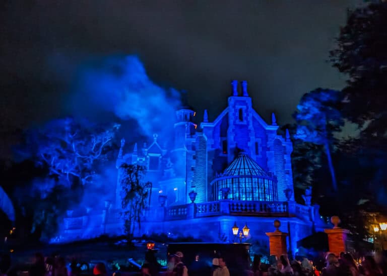 The Haunted Mansion at Disney World glows with eerie blue lighting at night as thick clouds of fog drift above the Gothic style building. Guests gather in front of the attraction beneath the dark sky while surrounding trees and warm lanterns add to the spooky atmosphere.