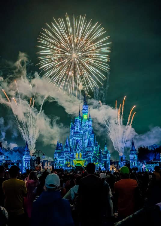 A spectacular burst of golden fireworks lights up the night sky above Cinderella Castle at Magic Kingdom in Walt Disney World. The castle glows with blue and green projections as fireworks and clouds of smoke surround its towers, while a large crowd of guests watches the nighttime fireworks show.