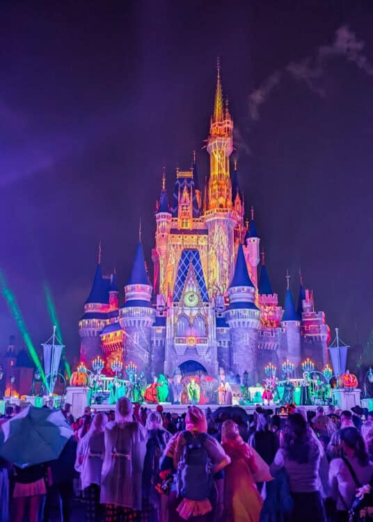Cinderella Castle at Magic Kingdom in Walt Disney World glows with orange, purple, and green Halloween projections during a nighttime stage show. Performers in colorful costumes dance on a stage decorated with glowing jack o lanterns and candelabras while a large crowd watches beneath the dark sky.