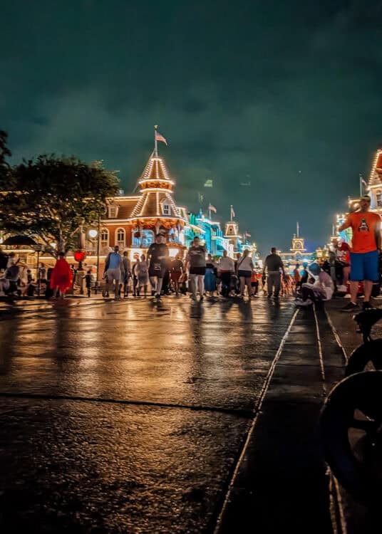 Guests walk along Main Street U.S.A. at Magic Kingdom in Walt Disney World at night, surrounded by Victorian style storefronts outlined in glowing lights. The wet pavement reflects the warm lights of the buildings beneath a dark, cloudy sky.