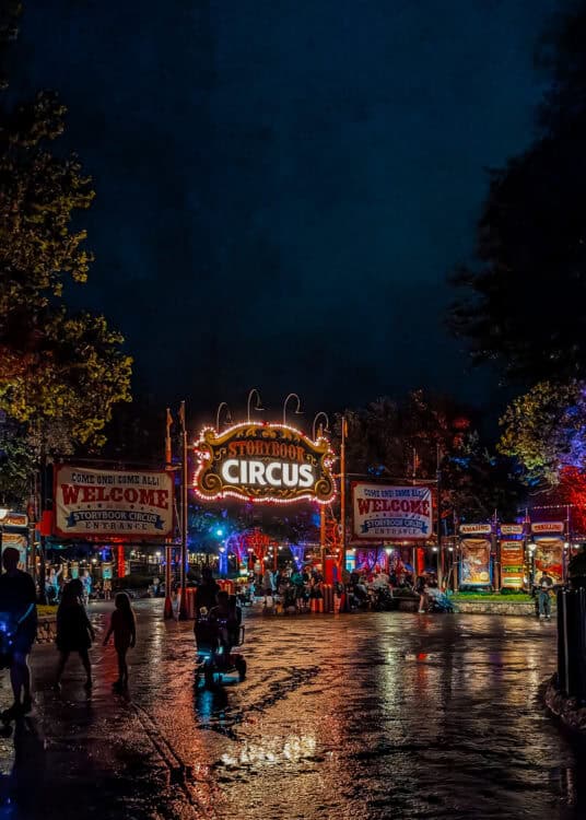 The Storybook Circus entrance at Magic Kingdom in Walt Disney World glows with colorful carnival lights at night. The illuminated sign reads "Storybook Circus" above the entrance, while two large banners welcome guests with the words "Come One Come All. Welcome. Storybook Circus Entrance." Visitors walk along the wet pavement reflecting the bright lights of the circus themed area.